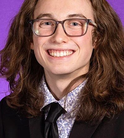 Zasha Griffin smiles in front of a purple painting. He has long, curly, brown hair and wears glasses, a bright purple floral shirt, a black tie and a black suit jacket.